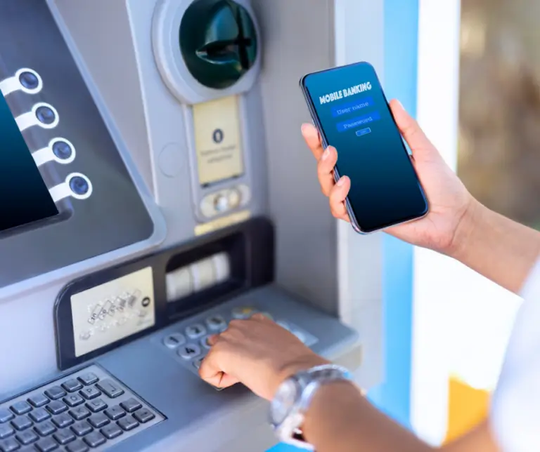 Hands interact with an ATM and a cell phone simultaneously. The person is using a mobile banking app. The image represents seamless banking and omni-channel banking.